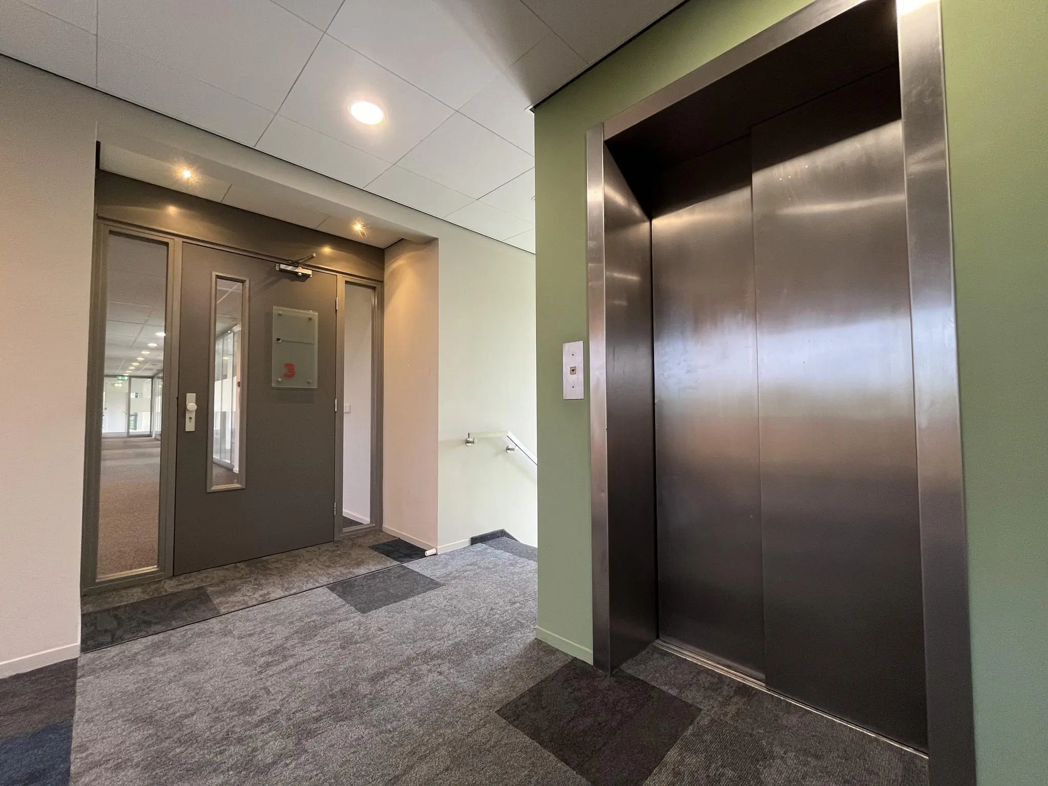 Hal met een gesloten liftdeur en een grijze toegangsdeur in een kantoorgebouw aan de Baarnsche Dijk.